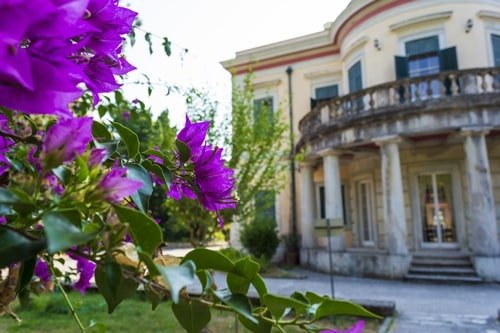 Außenansicht der hellen Villa mit Säulen im Eingangsbereich und blühendem Blumenstrauch.