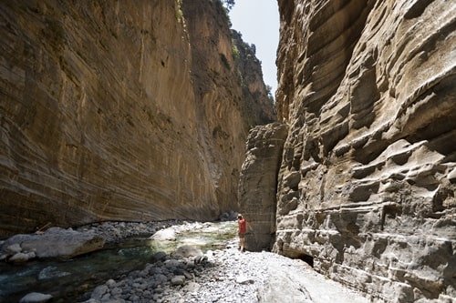 Eine Frau wandert durch die Schlucht von Samaria. Neben ihr ein Fluss und die bis zu 600 Meter hohen Bergwände der Schlucht.