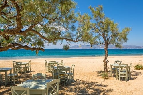 Holztische und Stühle stehen an Nadelbäumen am Plaka Beach auf Naxos