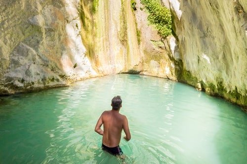 Mann in Badehose steht in natürlichem Wasserbecken vor kleinem Wasserfall